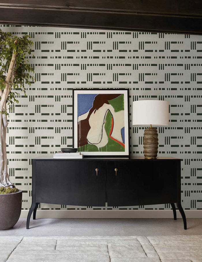 | The Artemis green and white dash-and-bar motif patterned wallpaper is in a room with a black wooden console table, an abstract colorful wall art, and a brown ribbed lamp