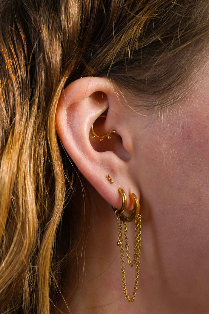 Close up of a model's ear with multiple earrings, including an earring charm featuring a length of metal chain with a metal ball dangling at the bottom. The charm is made from ASTM F136 Implant Grade titanium, and finished with 18K Gold PVD Plating