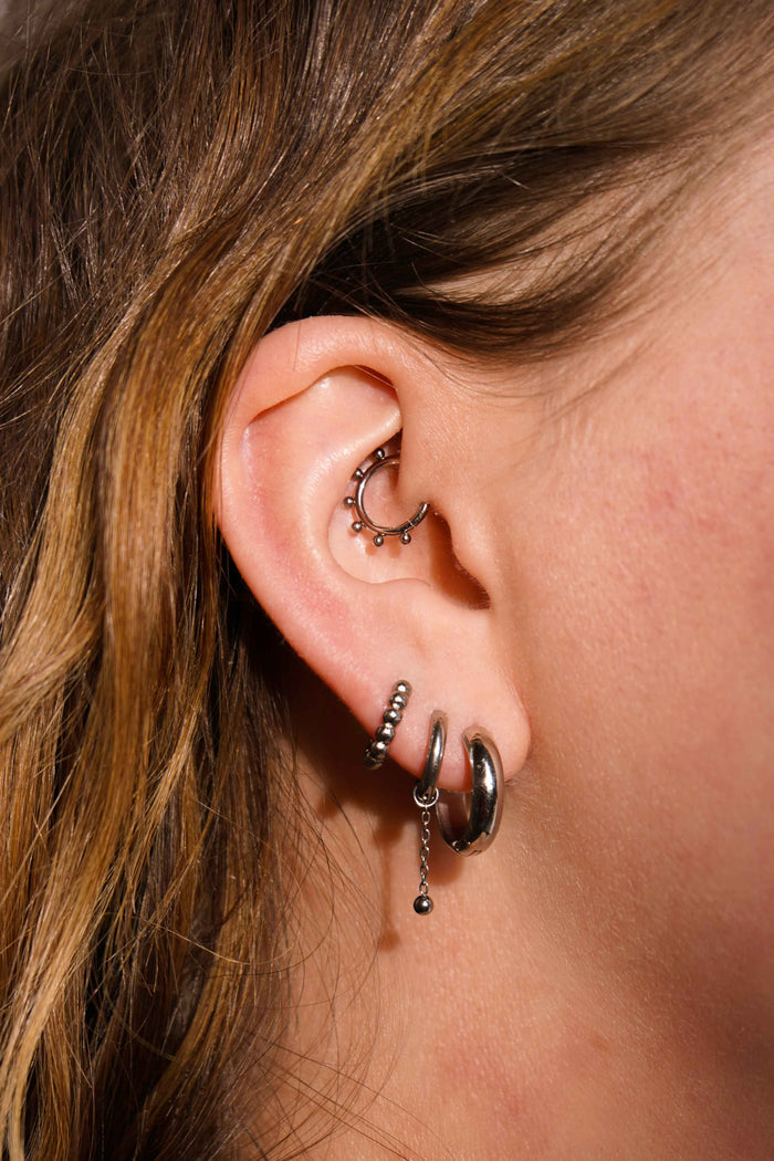 Close up of a model's ear with multiple earrings, including an earring charm featuring a length of metal chain with a metal ball dangling at the bottom. The charm is made from ASTM F136 Implant Grade titanium.