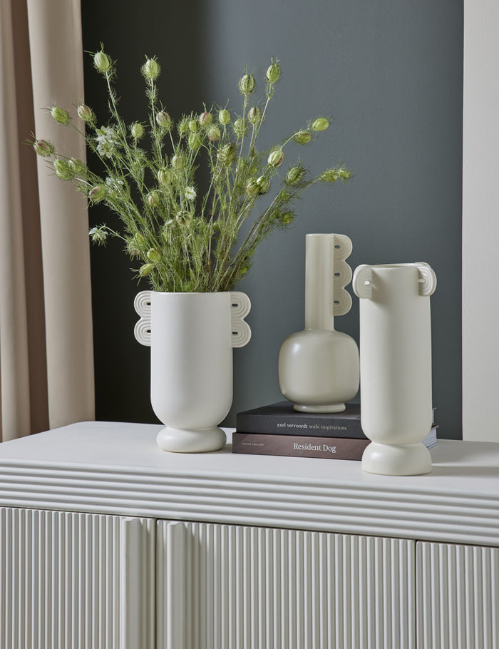 | The Marguerite matte white ceramic vase with a grooved neck sits with other white vases atop a textured white sideboard