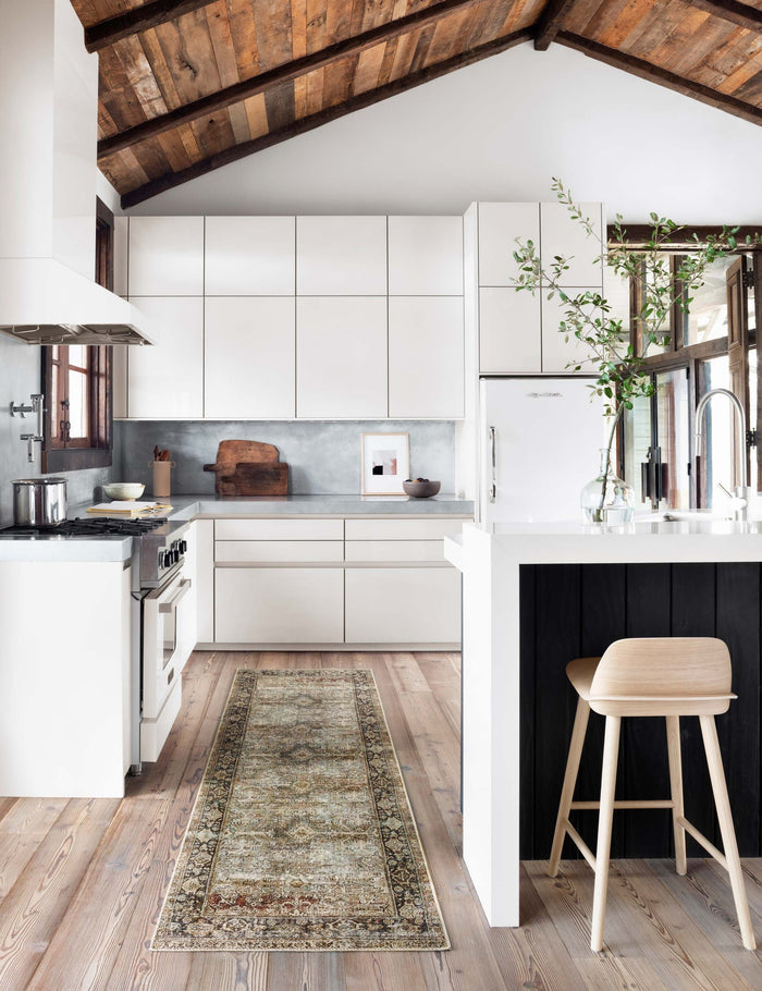 #size::2-6--x-12-0- #size::2-6--x-7-6- #size::2-6--x-9-6- #color::olive-and-charcoal | The Dacion distressed dark-toned persian rug in its runner size lays in a kitchen with white cabinetry, a natural wooden bar stool, and hardwood floors