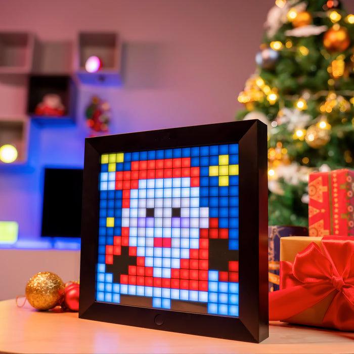 Divoom Pixoo 16x16 pixel art LED display, a Santa face with stars beside Christmas presents and a lit tree, smartphone app controlled, holiday gaming room decor