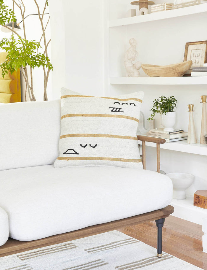 #style::square | The Iconic ivory stripe square pillow sits on a white sofa with a wood frame with a decor filled shelf in the background