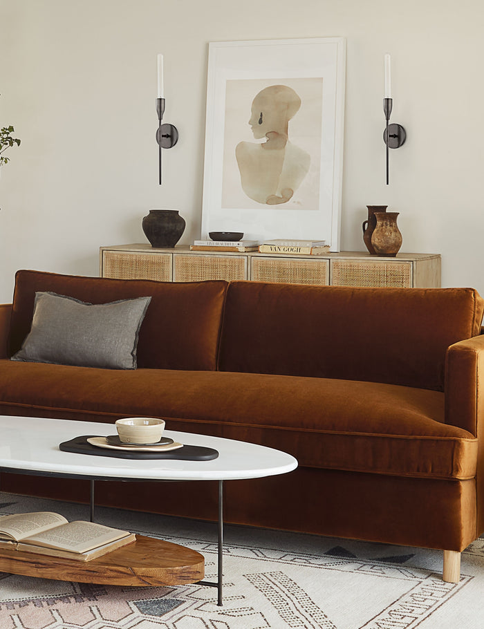 | Two Kama sleek, industrial black sconces with an aged bronze finish sits in a living room with a portrait painting in between them, a cognac velvet sofa, and a woven sideboard