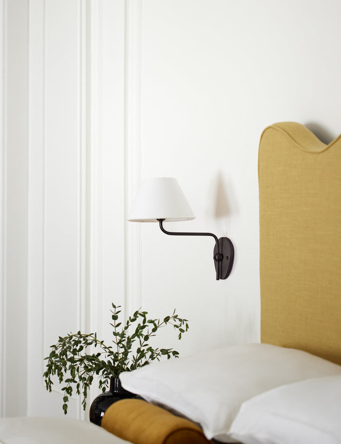 #color::white #finish::bronze | The Magdalene black single sconce is mounted to the right of a golden linen bed on a white wall