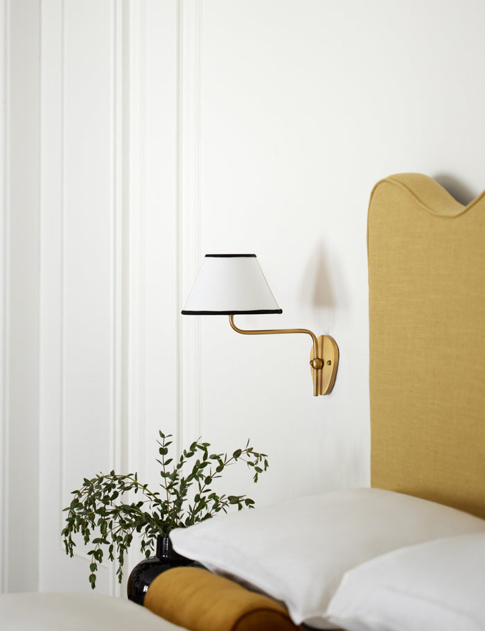#color::black-trim #finish::brass | The Magdalene brass single sconce is mounted to the right of a golden linen bed on a white wall