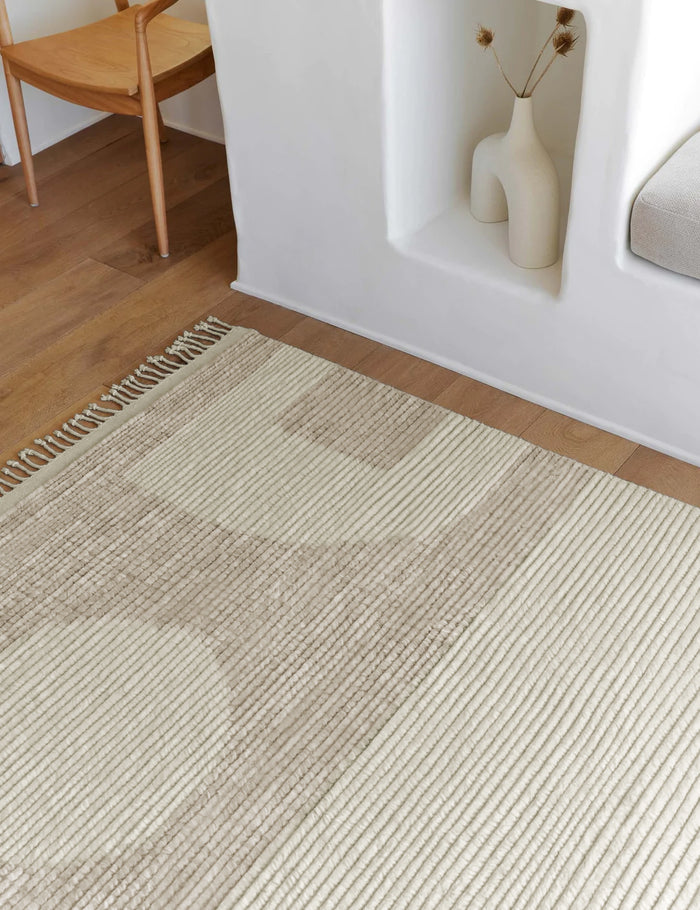 #size::2-6--x-8- #size::6--x-9- #size::8--x-10- #size::9--x-12- #size::10--x-14- #size::12--x-15- | Close-up of the Nomad neutral-toned geometric floor rug by Élan Byrd with subtle ribbed design in a room with a sculptural white vase and a neutral wooden dining table atop hardwood floors