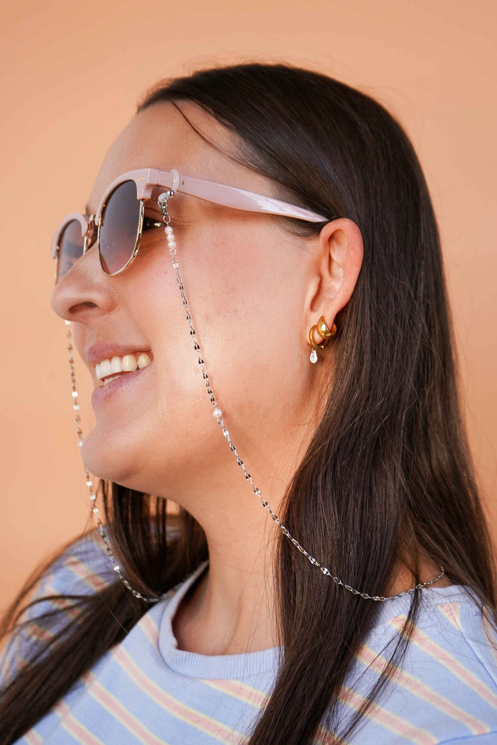 Woman wearing sunglasses with a silver chain, smiling against a beige background.