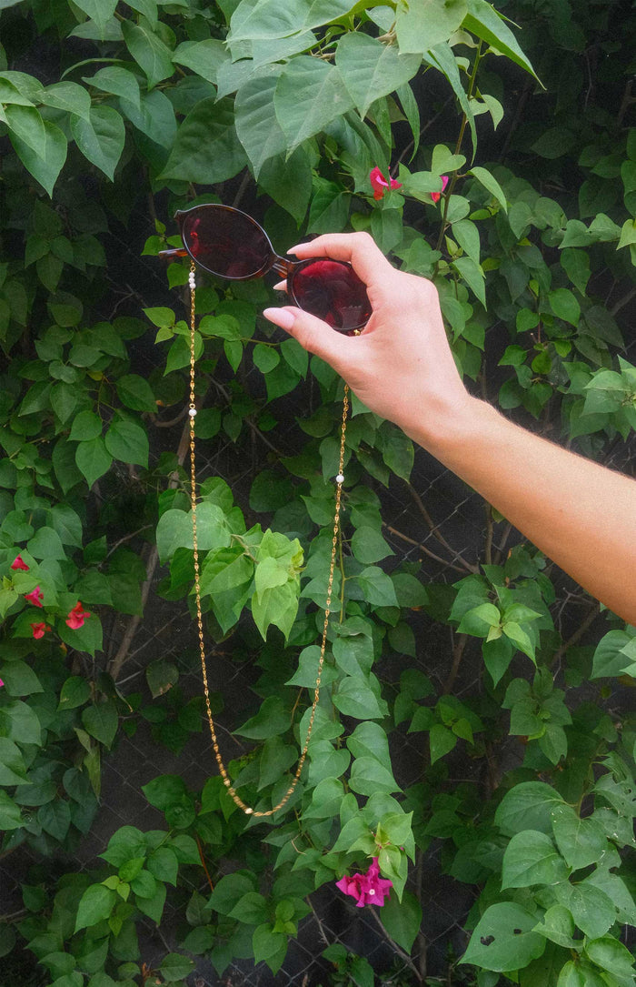 Hand holding sunglasses with a chain against a green leafy background
