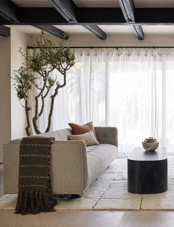 | The Pismo dark gray pure wool throw blanket with fringed ends lays on a gray linen sofa in a living room with a cream plush rug