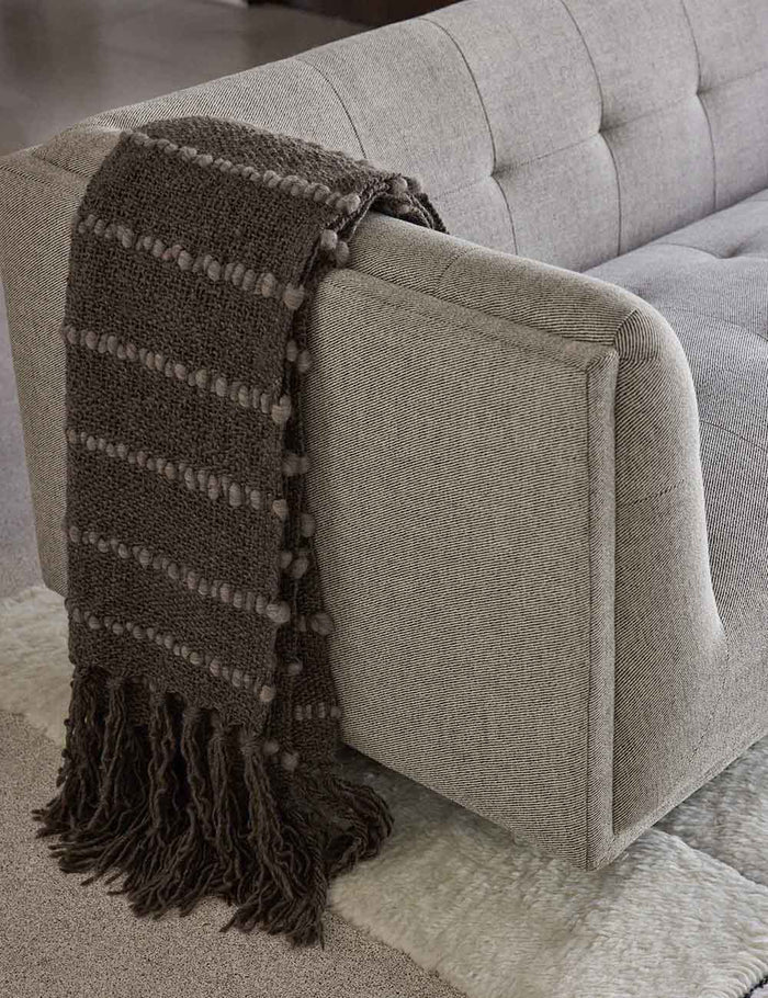 | The Pismo dark gray pure wool throw blanket with fringed ends lays on a gray linen sofa in a living room with a cream plush rug