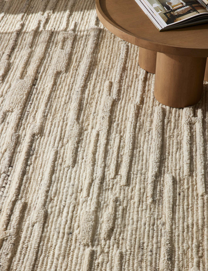 #color::neutral #size::2--x-3- #size::2-6--x-8- #size::5--x-7-6- #size::8--x-10- #size::9--x-12- #size::10--x-14- | The Rizzoli natural rug sits on a chevron wooden floor under a round wooden coffee table with cylindrical legs