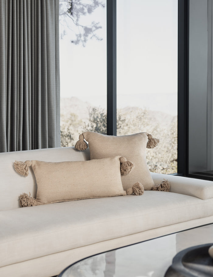 #color::terracotta #size::12--x-24- | Sami natural lumbar throw pillow with pom poms sits on a white linen couch