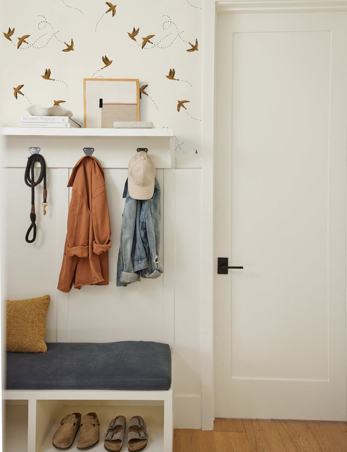 #color::rust | The Sparrow brown wallpaper by Rylee and Cru is in an entryway aboved an accented wall with a white coat rack and a blue cushioned bench.