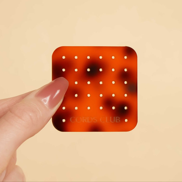 Tortoiseshell-patterned acrylic square with rounded corners and a grid pattern of little circle cutouts, held by a hand against a beige background.